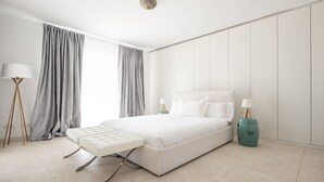 A modern bedroom with a large bed, bedside tables, a bedside lamp, and a view of the outside through sheer curtains. - Buccara Casa Bruno (Marbella)