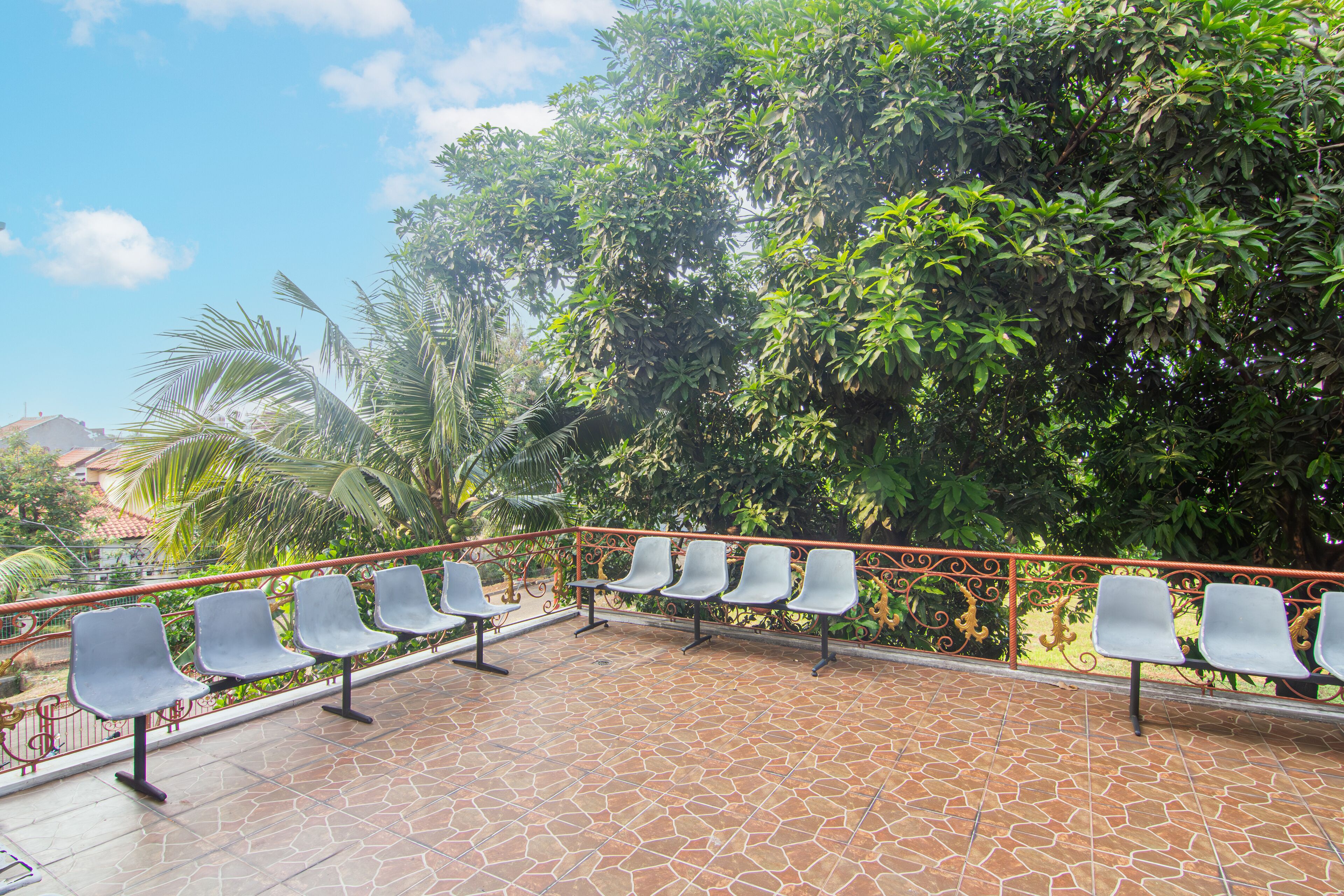 A terrace with a row of empty chairs and a tiled floor, surrounded by lush greenery and a metal railing.