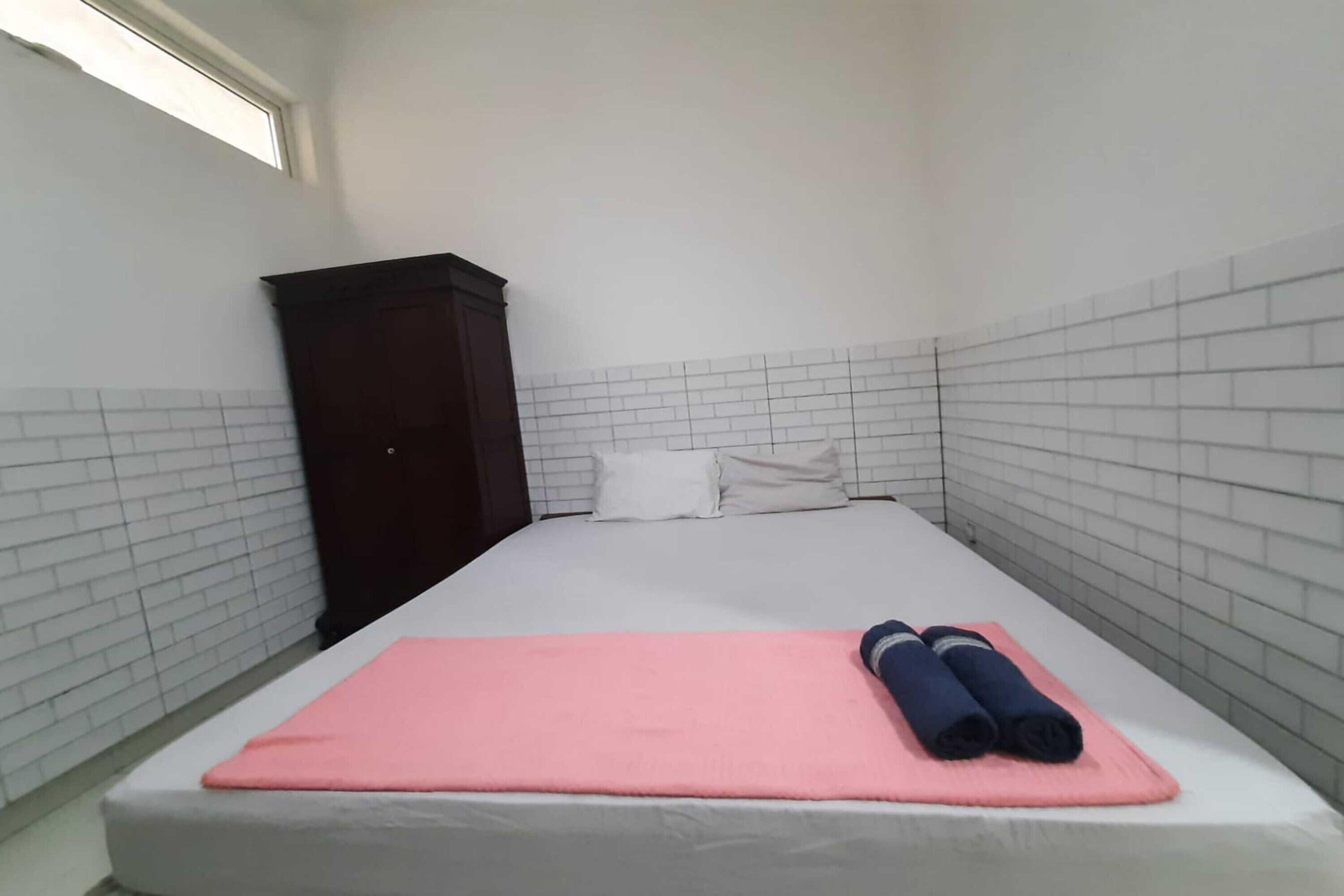 A small, narrow room with a bed, a pink blanket, and rolled towels.