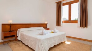 A neatly made bed with a white bedspread, two bedside tables with lamps, a window with brown curtains, and a wooden headboard.