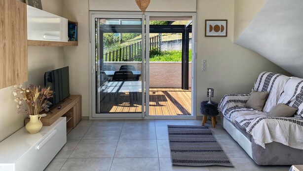 Una sala moderna con un sofá, un televisor, un estante de madera, un jarrón con flores secas y un ventanal corredero que da a un patio exterior.