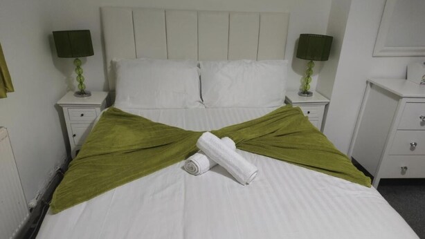 Een net opgemaakt bed met witte lakens, twee groene plaids en een opgerolde witte handdoek. Er staan twee nachtlampjes met groene lampenkappen op witte nachtkastjes.