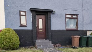 Een gebouw met twee verdiepingen, met een donkerbruine deur en ramen, een stenen pad dat naar de ingang leidt, en een kleine struik aan de linkerkant.