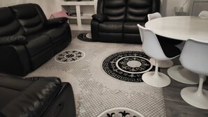 Een woonkamer met een zwarte leren bank, een witte eettafel met witte stoelen en een vloerkleed met een patroon.
