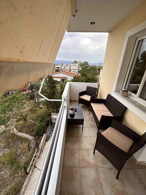 A balcony with wicker furniture, a small table, and a view of buildings and greenery. - VerGian Brand New Apt at Agioi Theodoroi (Loutraki-Agioi Theodoroi)
