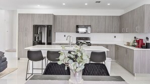 A modern kitchen with grey wood cabinetry, a central island with bar stools, and a dining table with a vase of greenery. - Avi Lux Stay III (Miami)