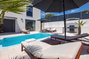 Un área de piscina moderna al aire libre con sillones de descanso, una estación de barbacoa y una zona de estar cubierta.
