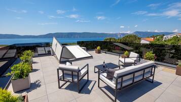 Terraço ao ar livre com assentos, vista para o mar e montanhas ao longe.