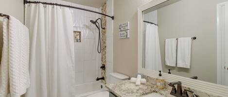 A bathroom with a shower curtain, toilet, sink with a marble countertop, and a large mirror.