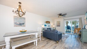 A spacious living area with a dining table, a sofa, a ceiling fan, and a view through a glass door. - Ocean House 2202 2 Bedroom Condo (Gulf Shores)