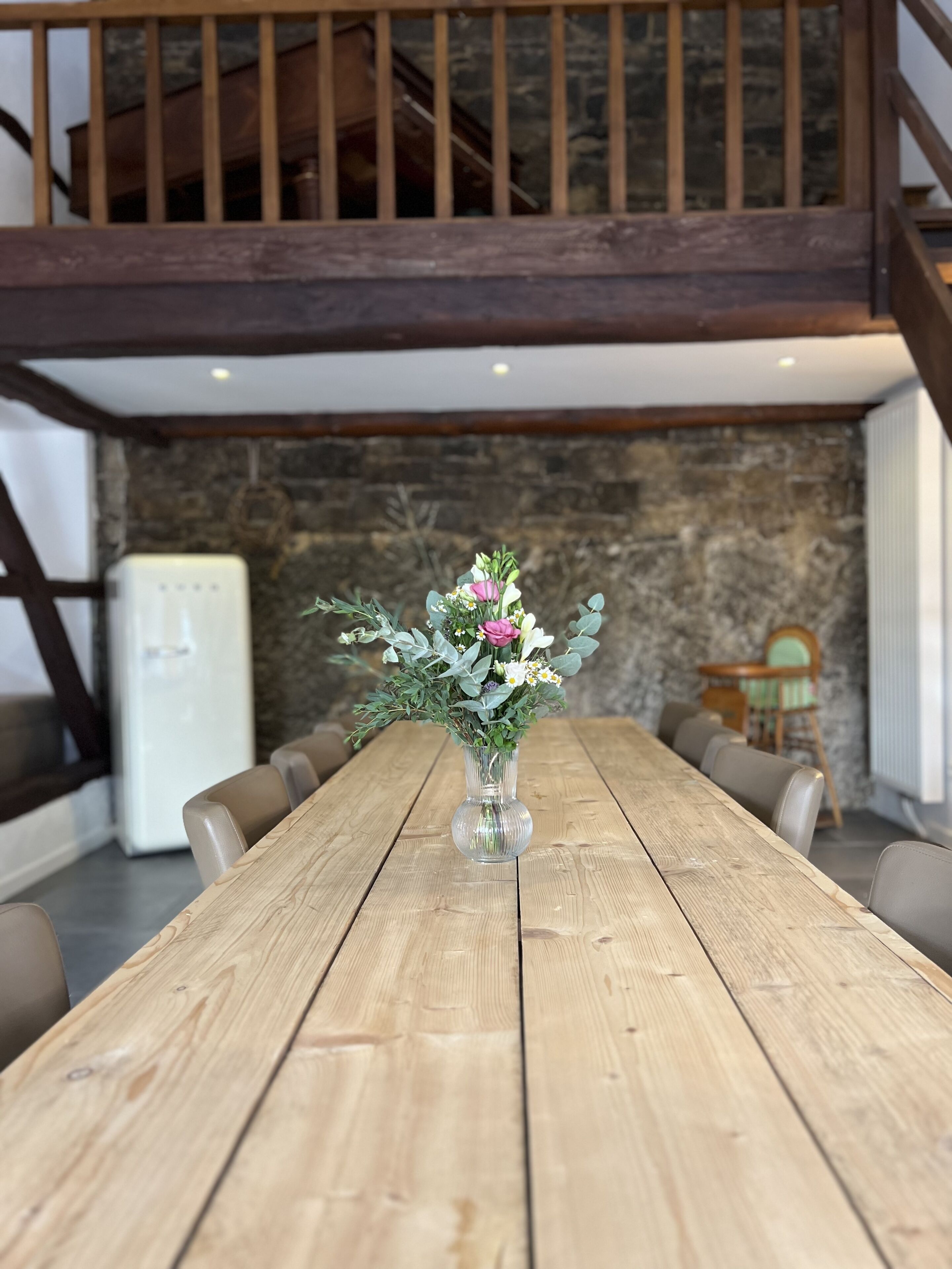 A wooden dining table with a vase of flowers in the center, set against a stone wall and a wooden staircase.