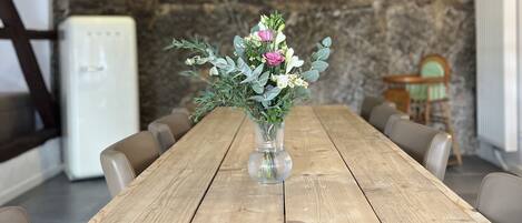 A wooden dining table with a vase of flowers in the center, set against a stone wall and a wooden staircase.