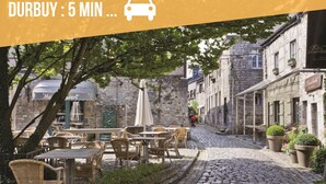 Une rue pavée avec des terrasses, des bâtiments en pierre et un symbole de voiture indiquant que Durbuy est accessible en 5 minutes en voiture.