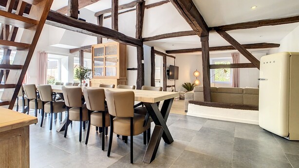 Un vaste espace de salle à manger avec une longue table et des chaises, un buffet en bois doté de portes en verre, et une cuisine moderne avec un réfrigérateur blanc.