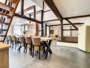 Un vaste espace de salle à manger avec une longue table et des chaises, un buffet en bois doté de portes en verre, et une cuisine moderne avec un réfrigérateur blanc.
