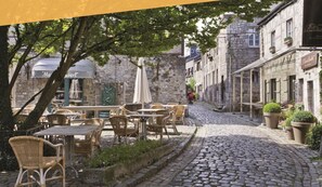 Une rue pavée avec des terrasses, des bâtiments en pierre et un symbole de voiture indiquant que Durbuy est accessible en 5 minutes en voiture.