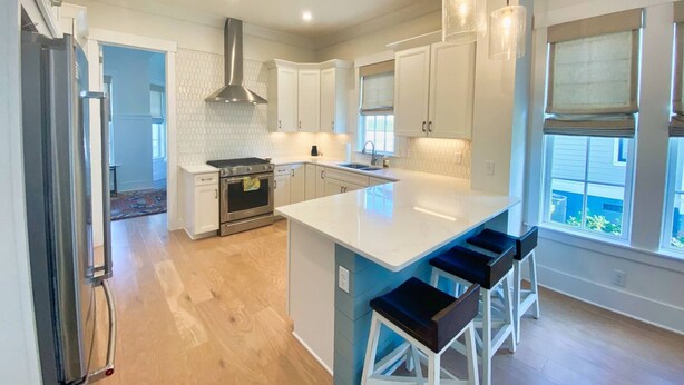 A modern kitchen with white cabinets, a stainless steel range hood, and a large island with bar stools. - Pebble Beach East Home with Bay Creek Amenities (Cape Charles)