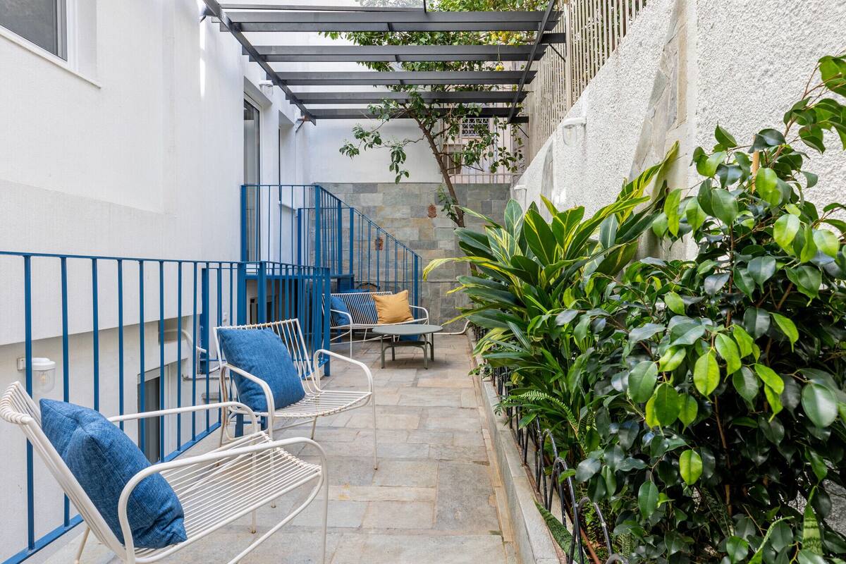 A narrow outdoor corridor with a metal railing, a blue bench with cushions, and lush green plants.