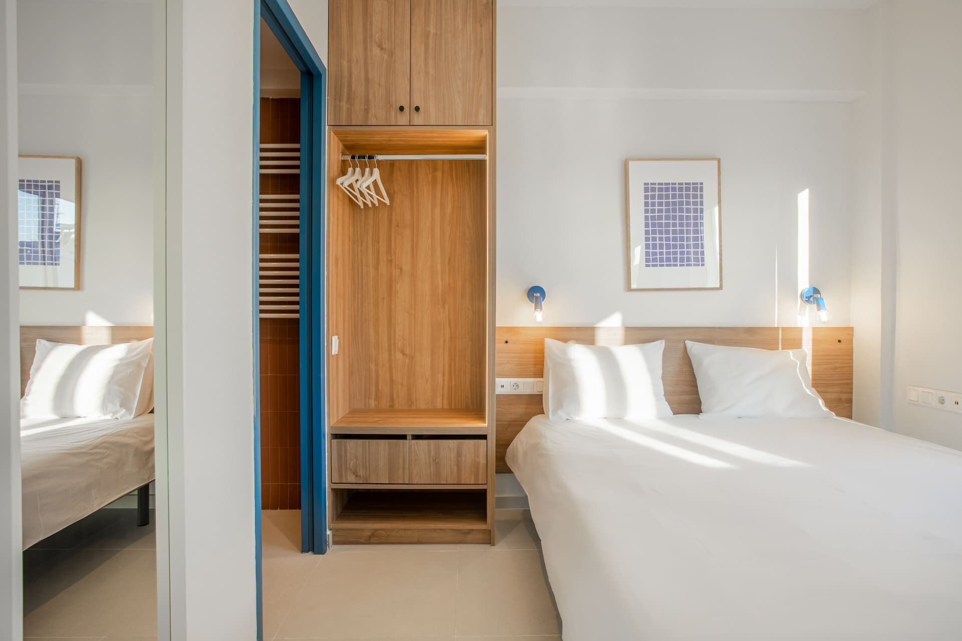 Een hotelkamer met een bed, een houten garderobe, een kledingkast met hangende kleding en een nachtkastje met een lamp.