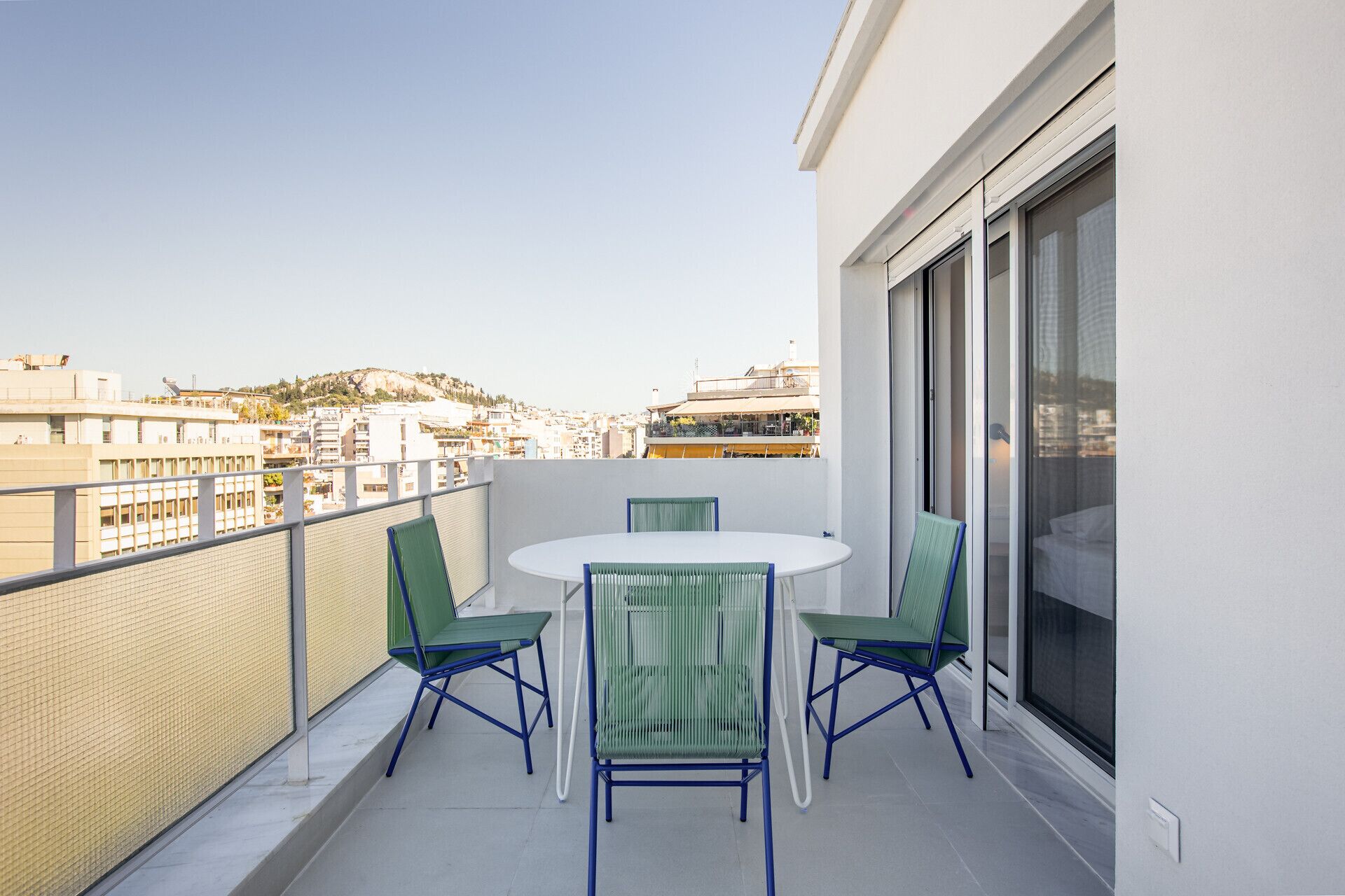 A balcony with a table and chairs, a view of buildings, and a large window.