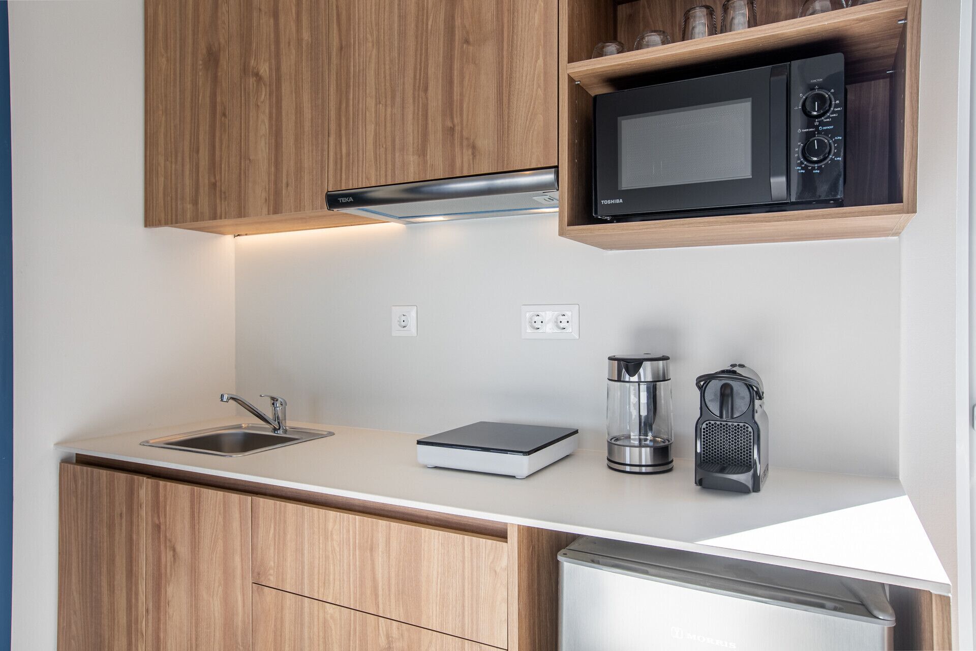 Cocina compacta con microondas, hervidor de agua y cafetera sobre la encimera, un lavabo y armarios de madera.