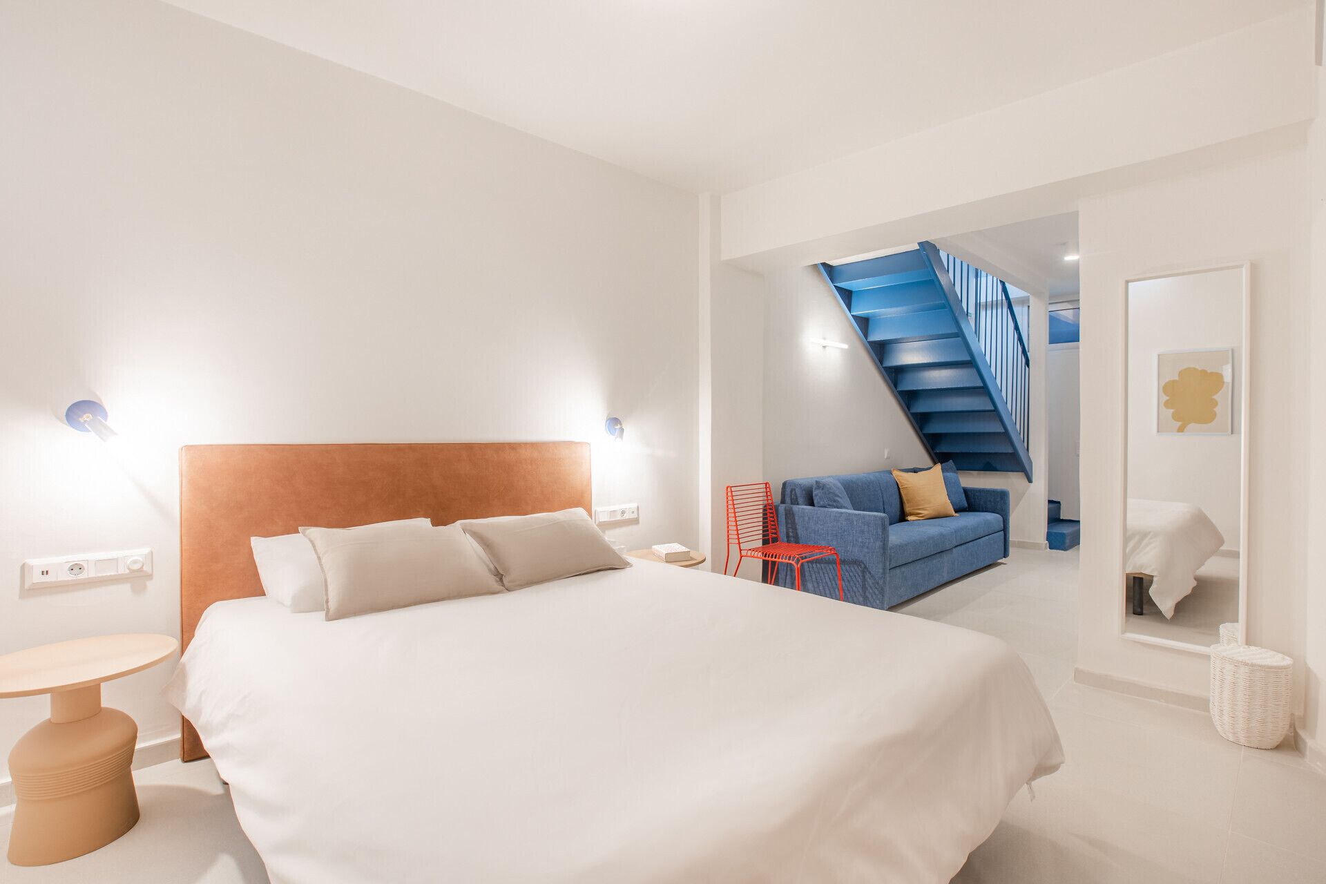 Ein modernes Hotelzimmer mit einem großen Bett, einem blauen Sofa, einem roten Sessel, einer Treppe und einem Spiegel.