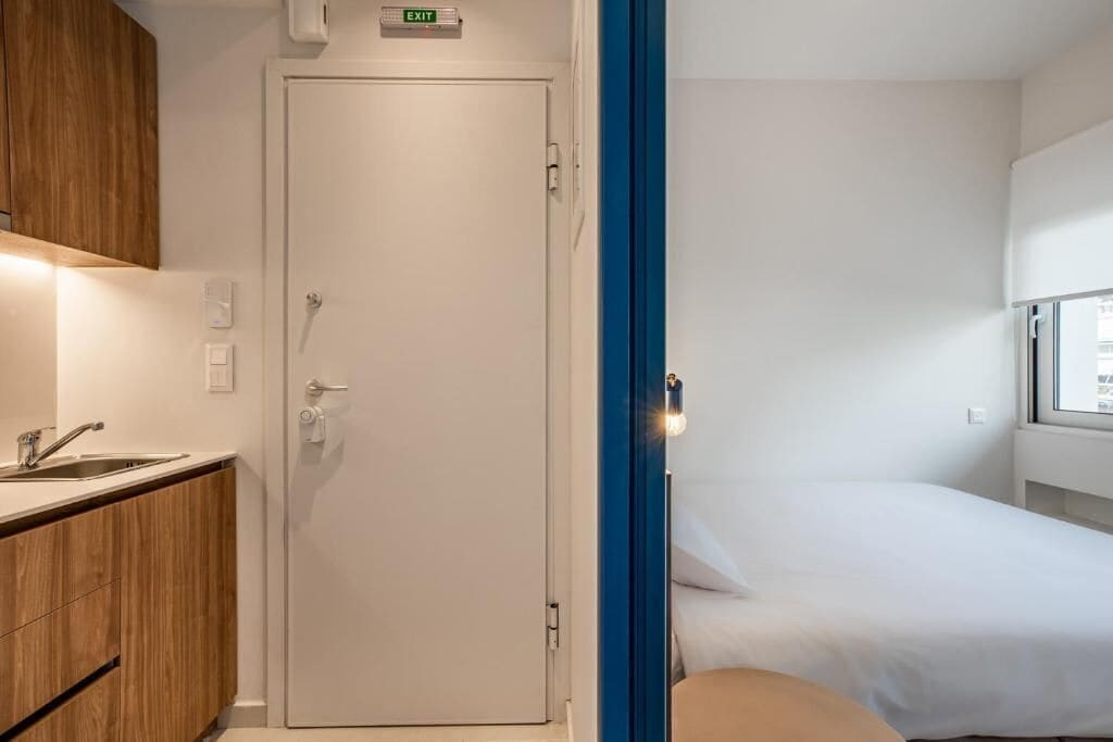 A hotel room with a bed, a window, a sink, and a door with an exit sign.