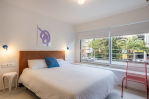 Habitación de hotel con cama, ventana con vista a palmeras y edificios, una silla roja y una lámpara fijada a la pared.