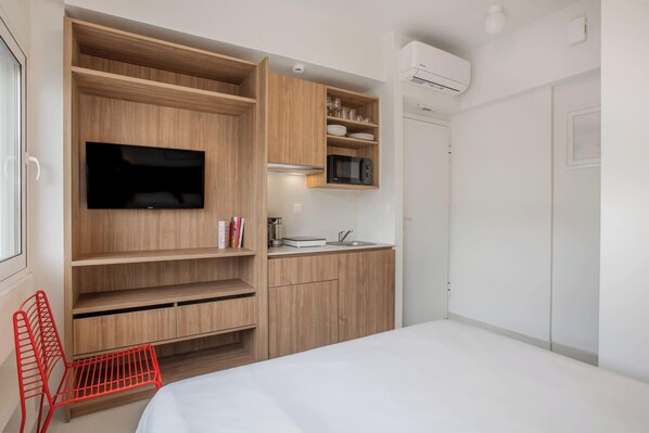 A compact room with a bed, built-in TV, kitchenette, and a red chair.