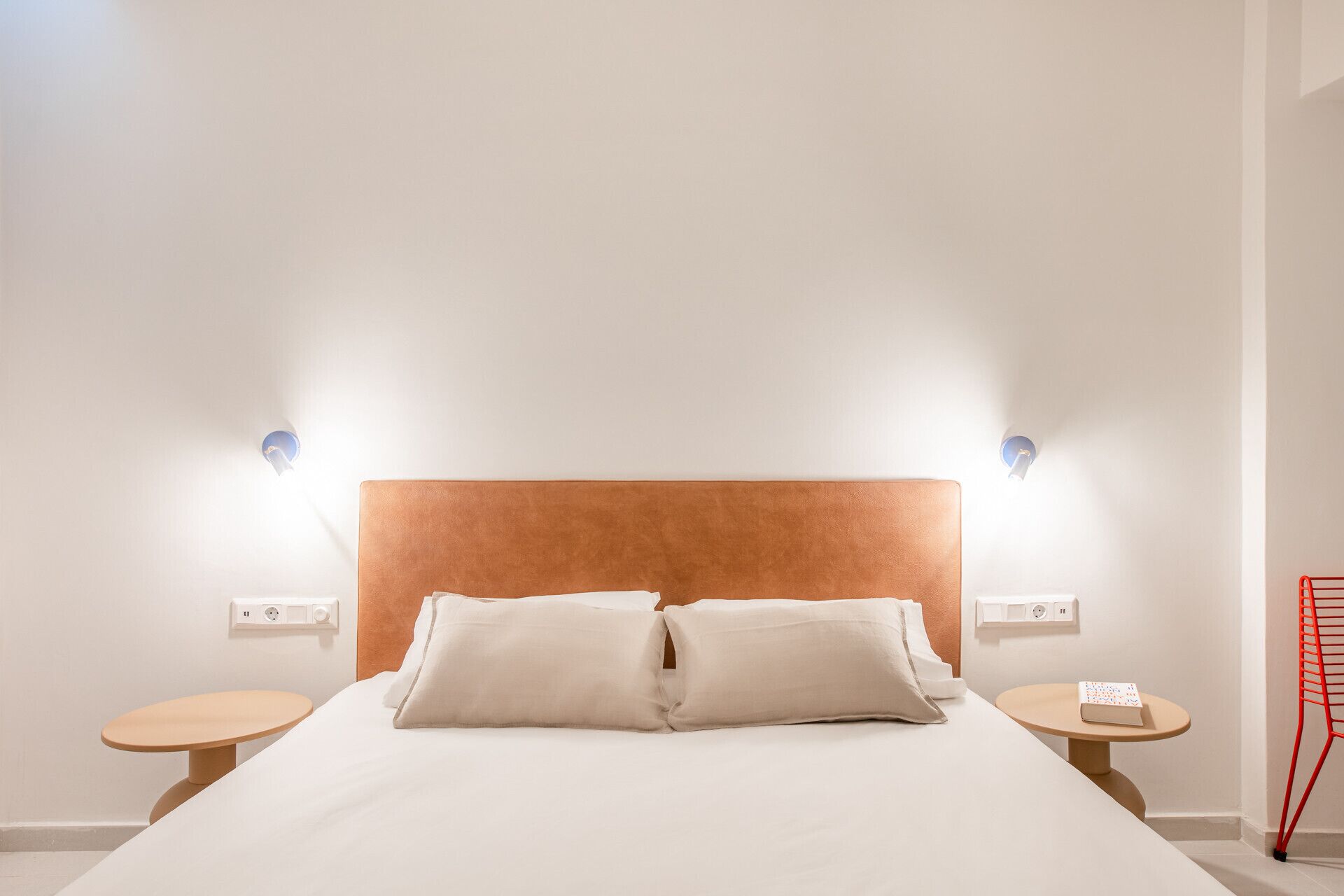 A hotel room with a large bed, two bedside tables, a red chair, and wall-mounted reading lights.