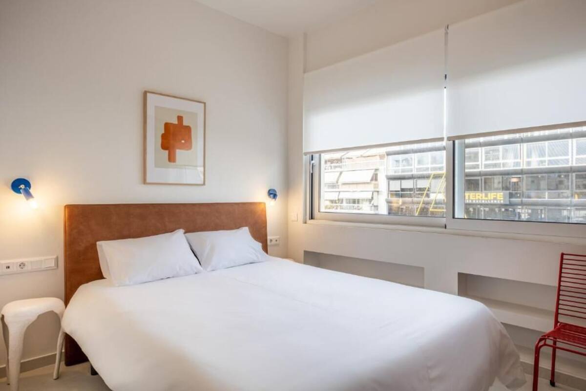 A hotel room with a large bed, a window with a view of a building, and a red chair.