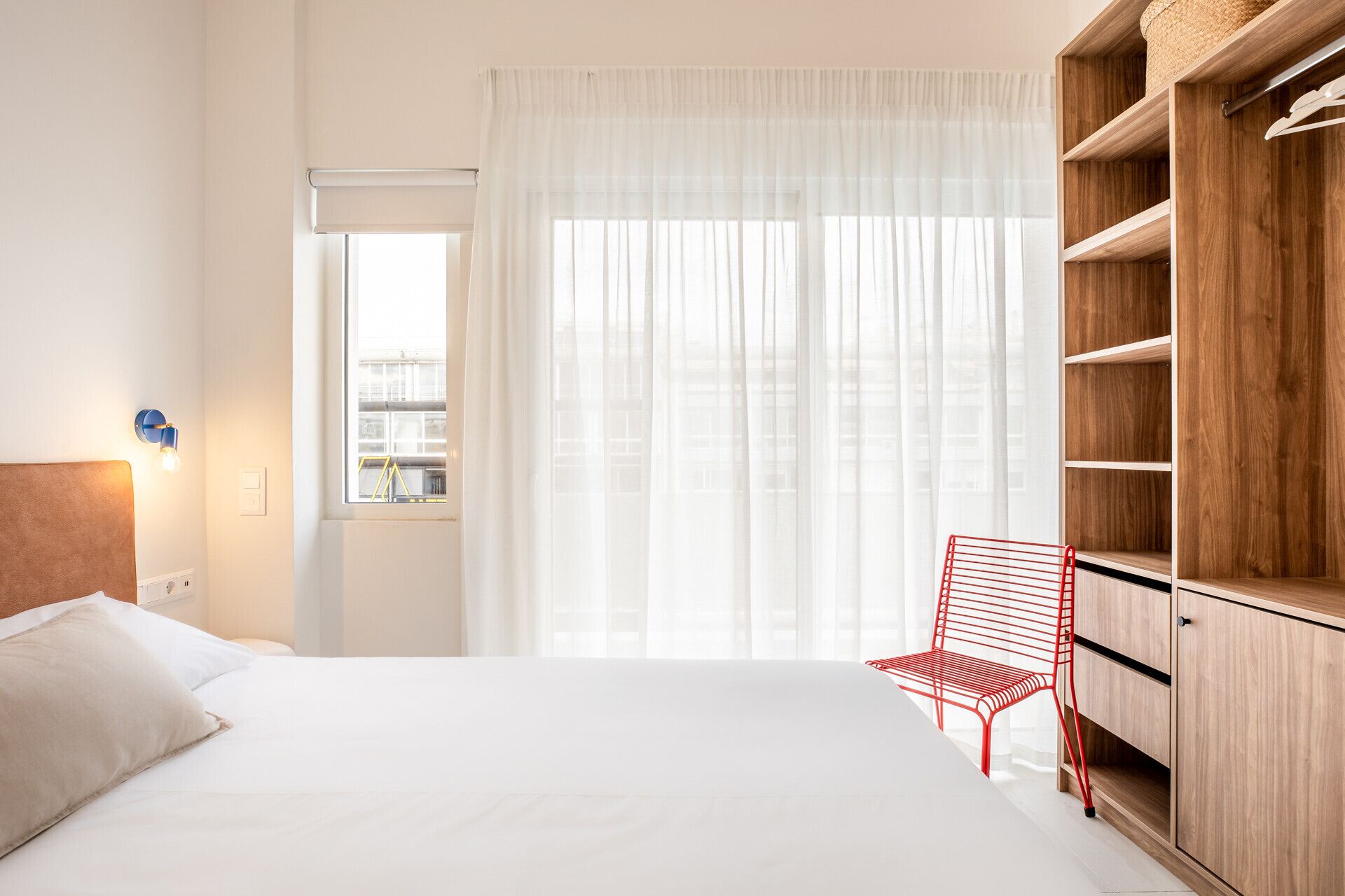 Et hotelværelse med en seng, en rød stol, et træskab og et vindue med lette gardiner.