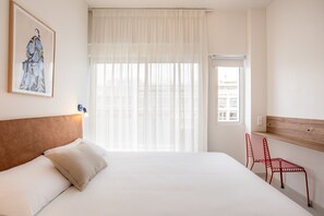 A hotel room with a bed, a window with sheer curtains, a red chair, and a framed picture on the wall.