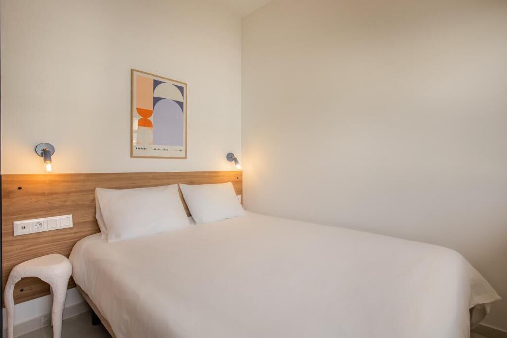 Una cama bien hecha con cabecera de madera, un taburete blanco pequeño y un cuadro abstracto colgado en la pared.