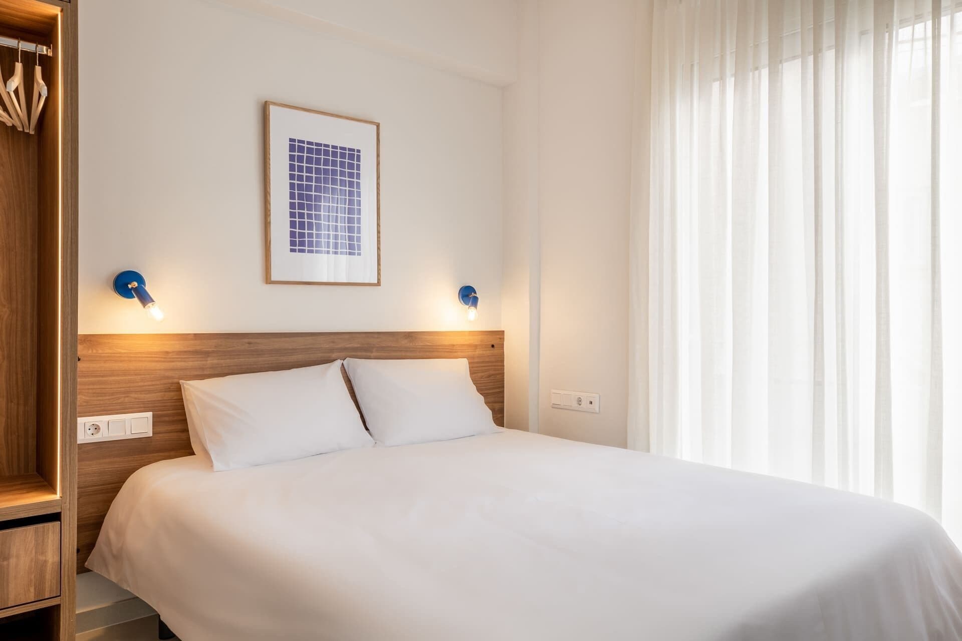 Quarto de hotel com cabeceira de madeira, uma obra de arte emoldurada, duas luminárias fixadas na parede e uma janela com cortinas translúcidas.
