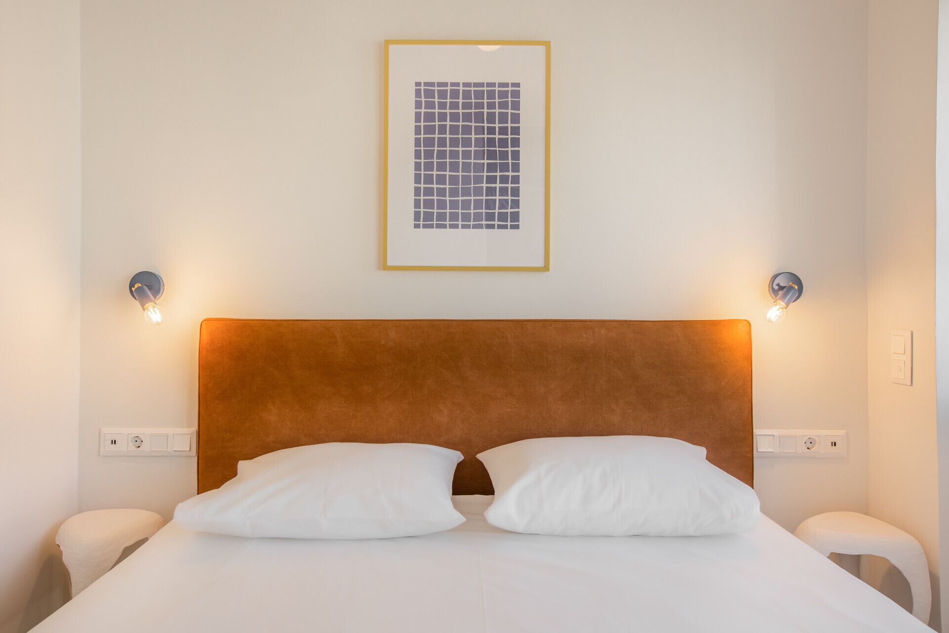 A hotel room with a brown headboard, white bedding, two bedside tables, and a framed picture on the wall.
