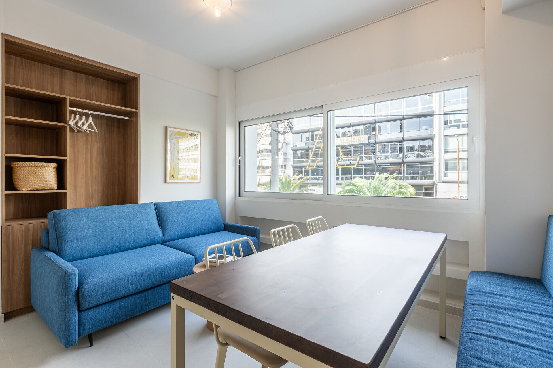 A modern room with a blue sofa, a dining table, and a large window with a view of buildings.