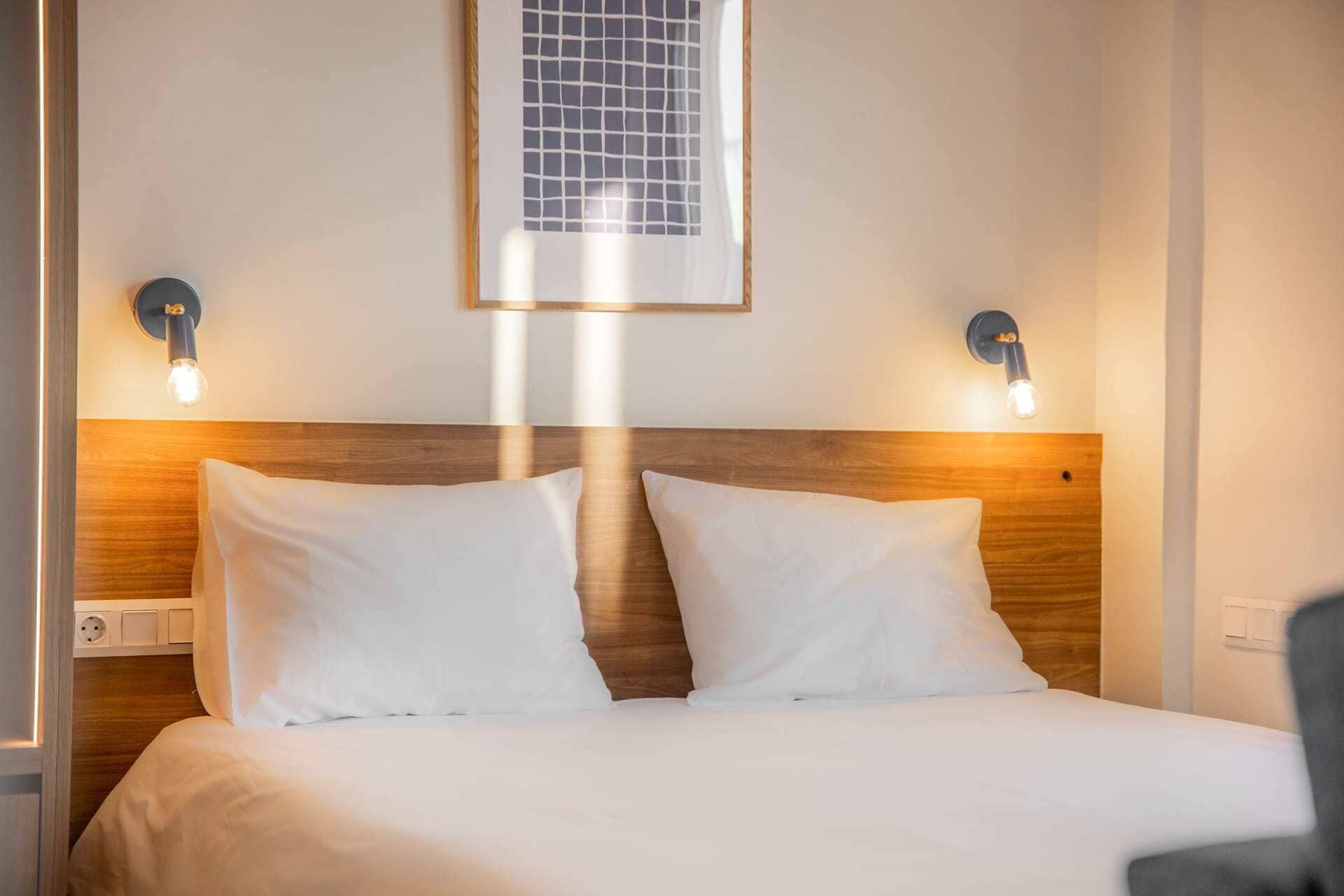 Een hotelkamer met een houten hoofdbord, twee witte kussens, een bed en twee wandlampen.