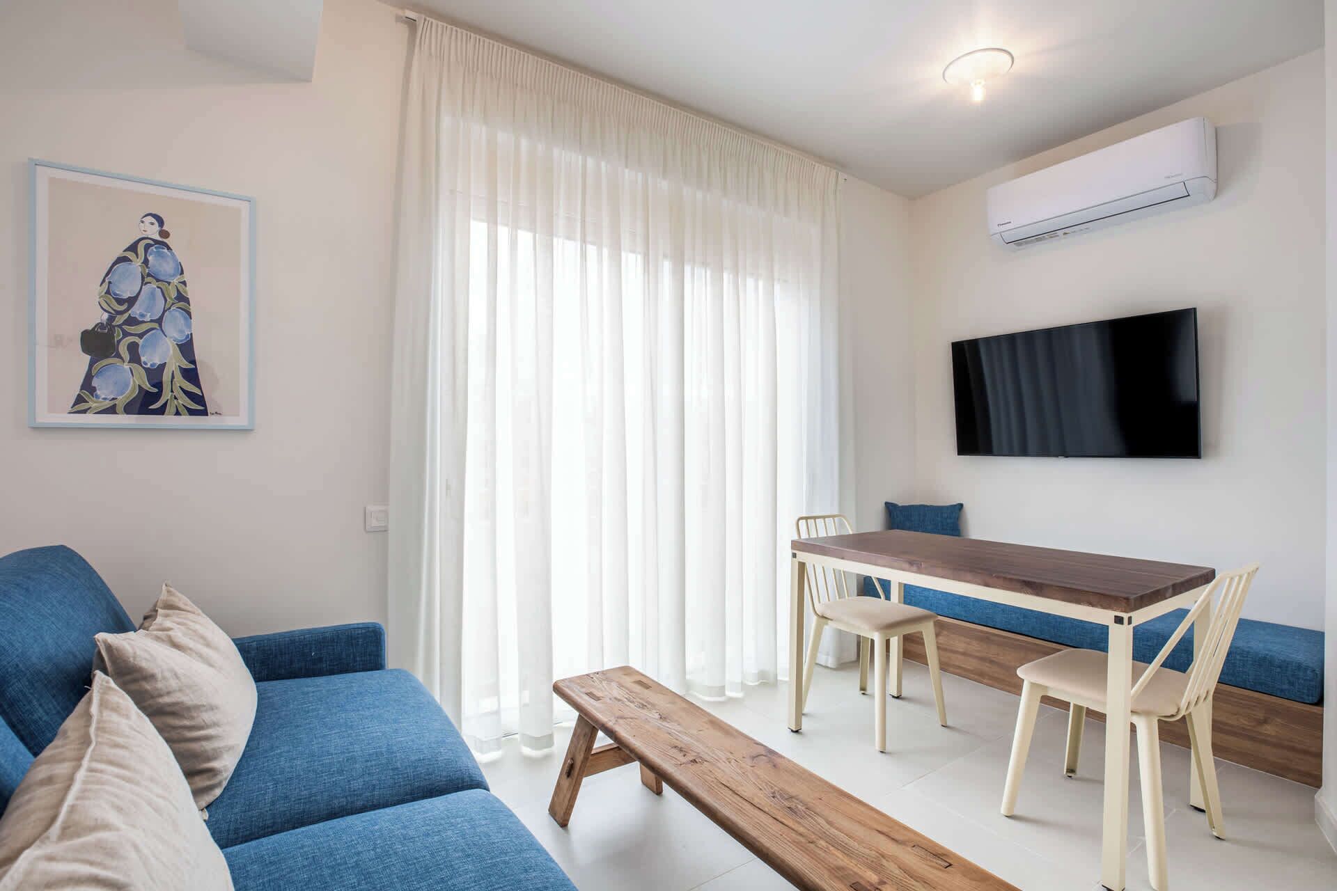 A living room with a sofa, a wooden bench, a dining table with chairs, a TV mounted on the wall, and a large window with sheer curtains.