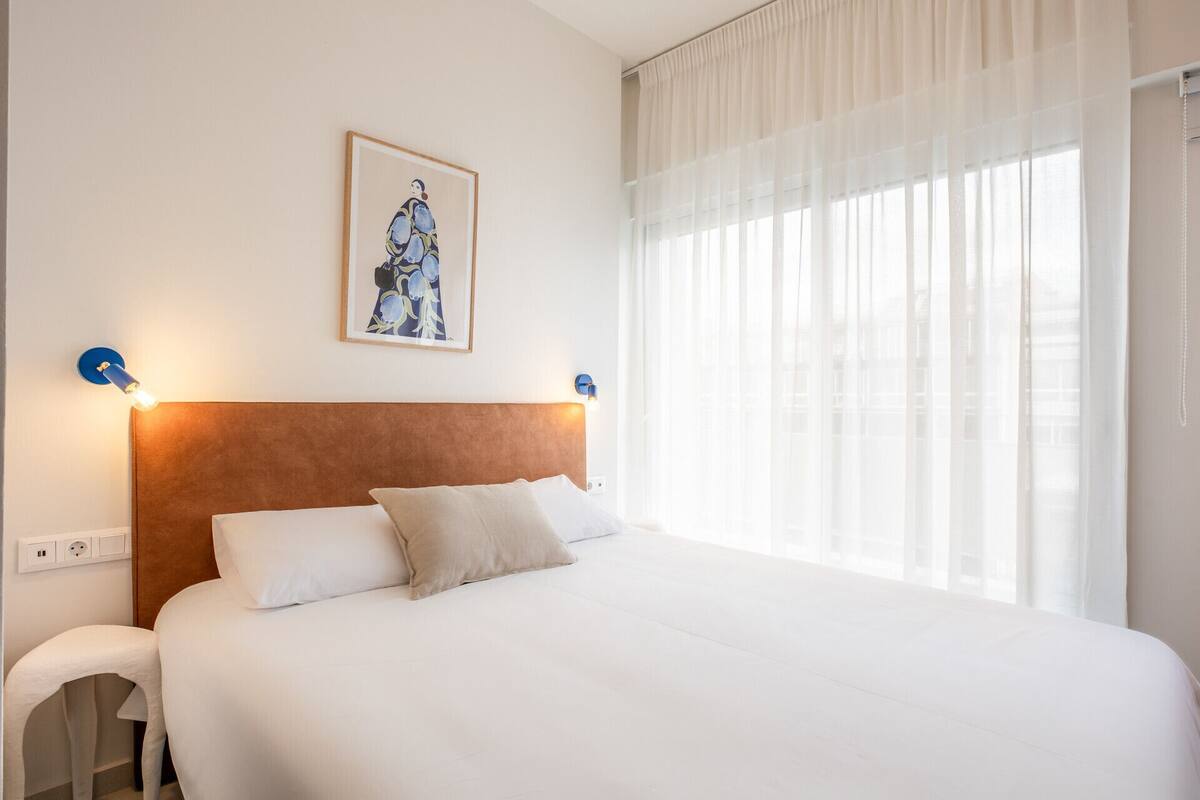 A hotel room with a large bed, a headboard, a wall-mounted light fixture, a power outlet, and a window with sheer curtains.