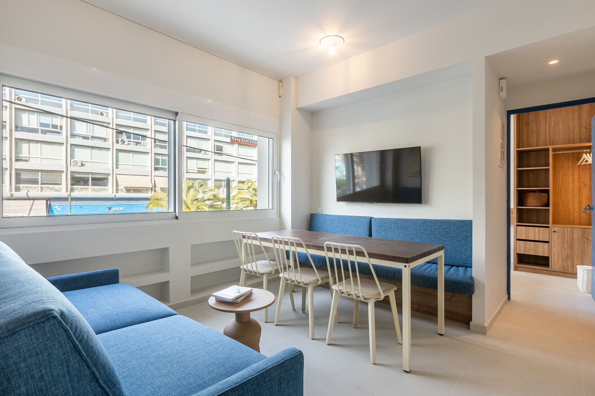 Ett modernt vardagsrum med en soffa, ett matbord, stolar, en tv och utsikt över en byggnad med en pool.