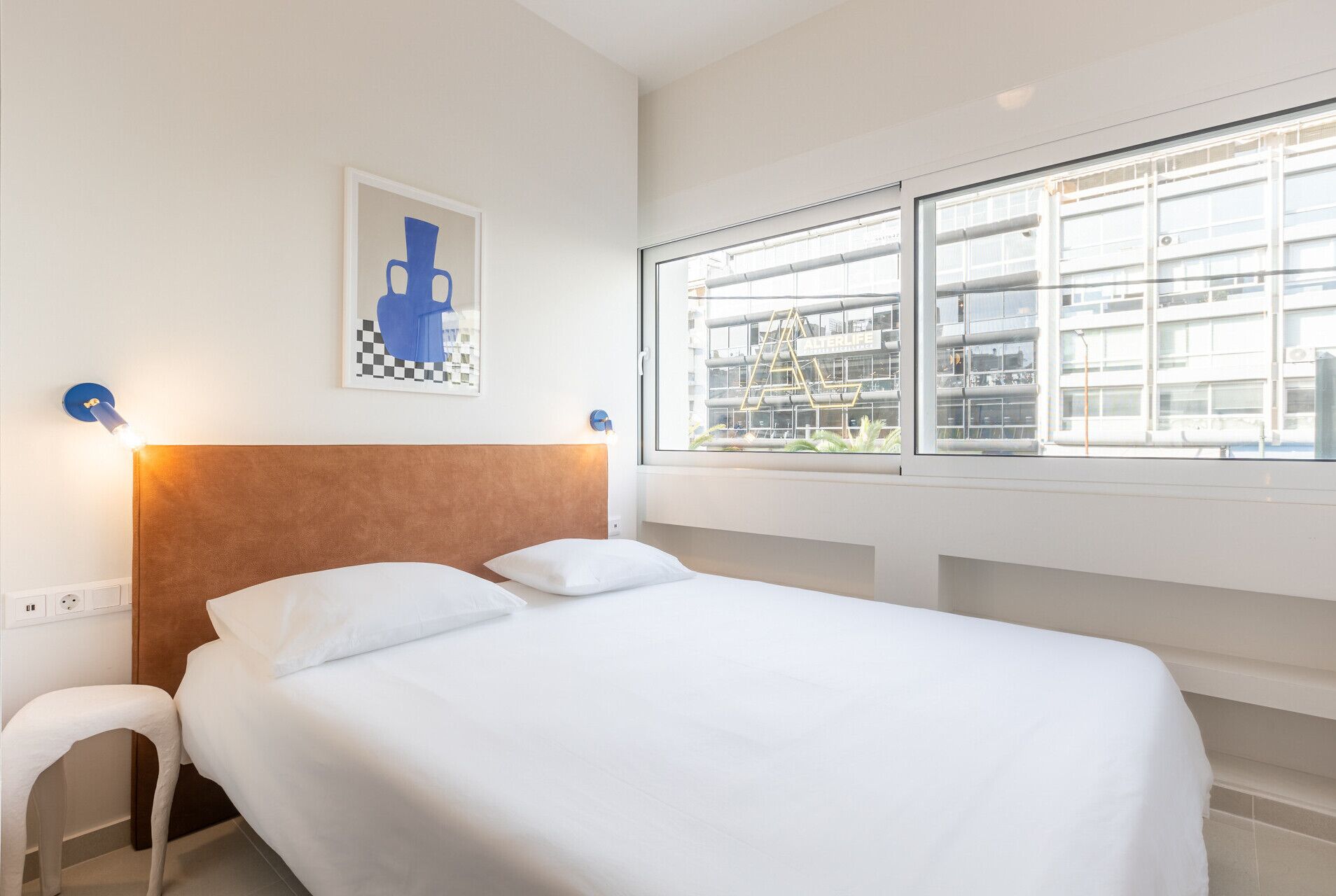 A hotel room with a bed, a window with a view of a building, and a wall-mounted artwork.