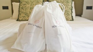 A bed with white linens, two white drawstring bags with 'RENT in PARIS.fr' printed on them, and a headboard with patterned pillows.
