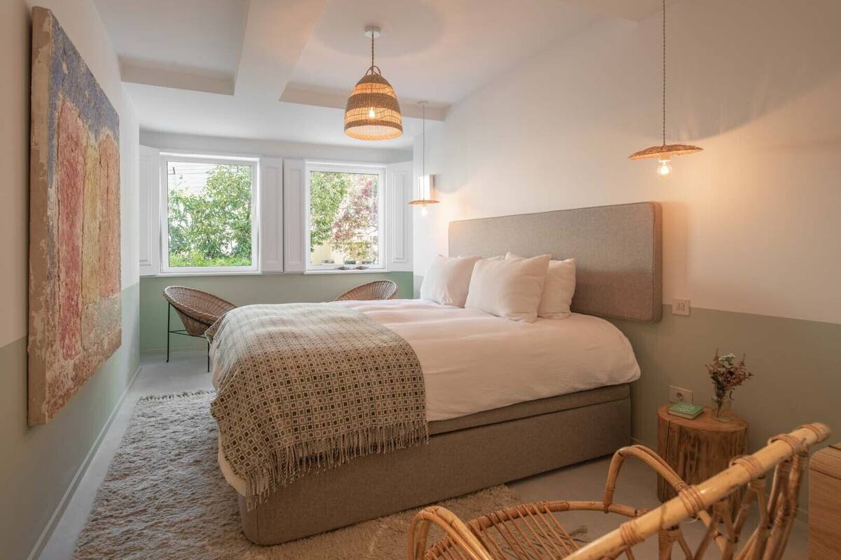 A bedroom with a bed, two hanging lights, a chair, a small wooden table, and a large painting on the wall.