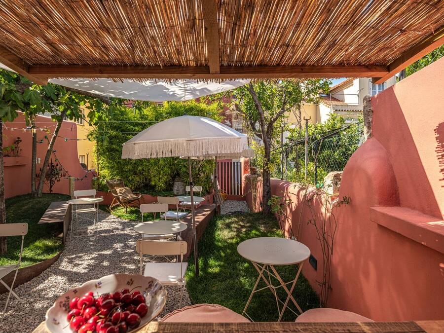An outdoor patio with a bamboo ceiling, white tables, and chairs, surrounded by greenery and a pink wall.