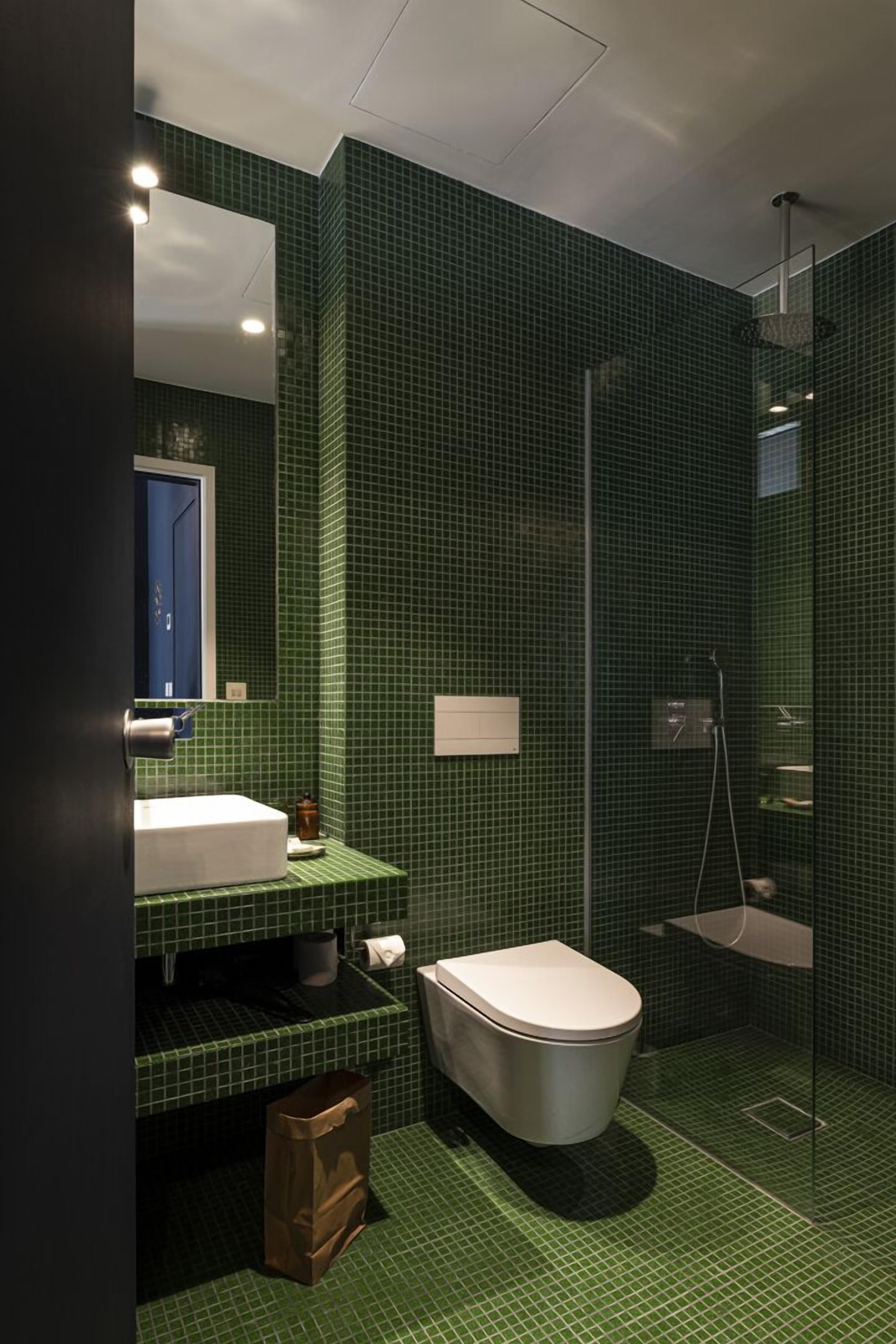 A bathroom with green tiled walls, a white sink, a toilet, and a glass-enclosed shower area.
