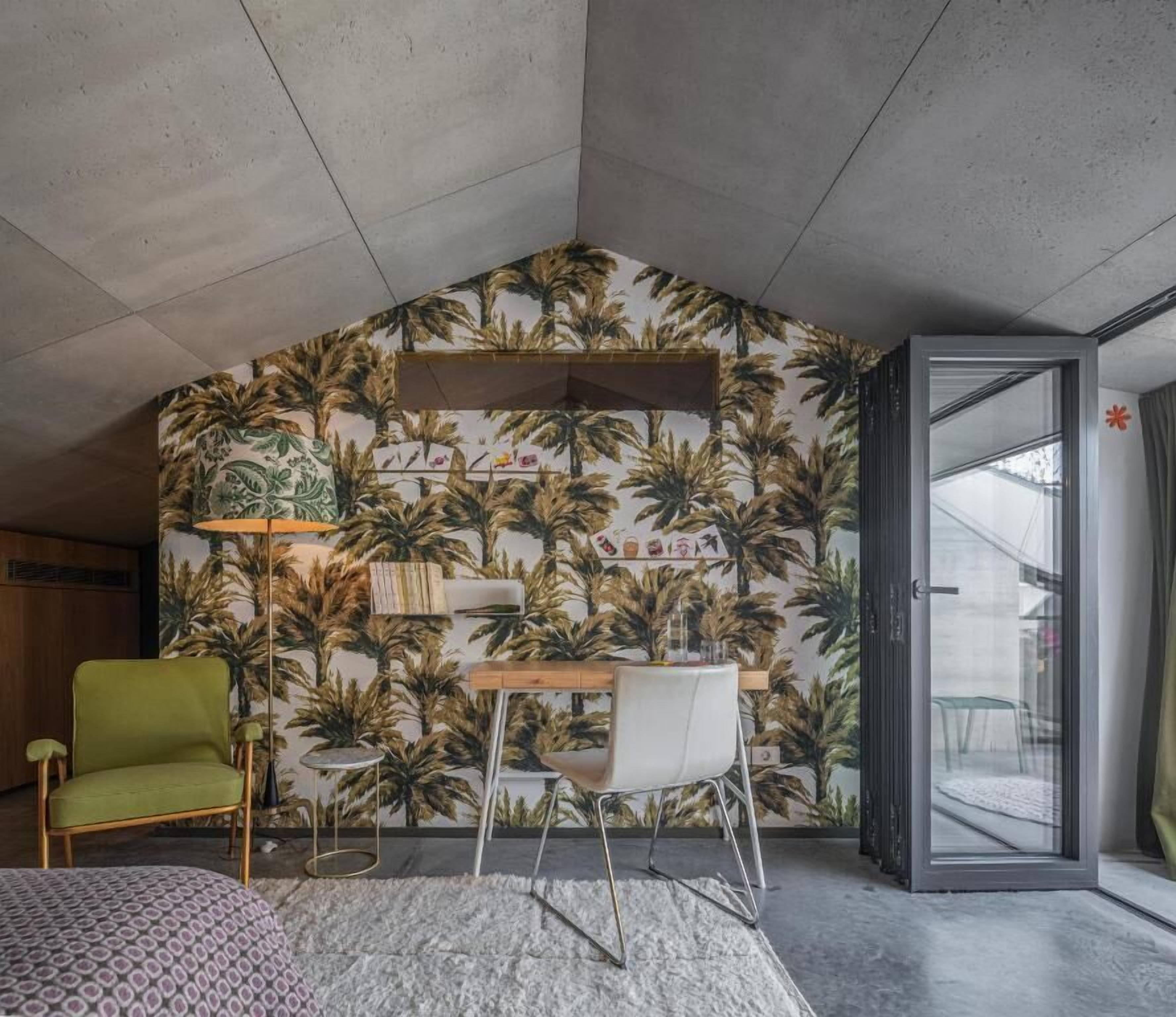 Une pièce moderne avec un papier peint aux motifs tropicaux, un fauteuil vert, une chaise blanche et une petite table en bois.