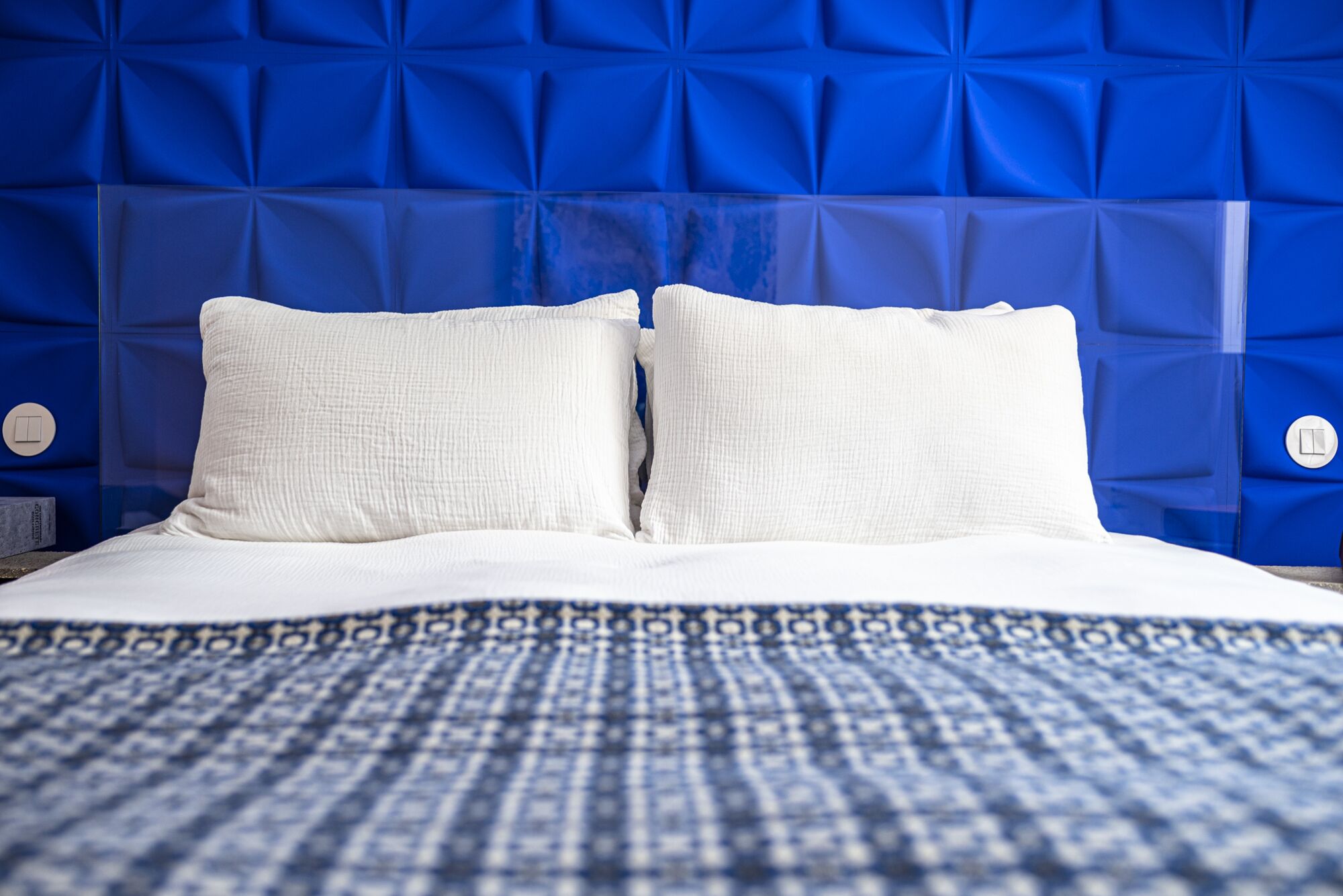 Un lit avec une literie blanche et un couvre-lit bleu à motifs, placé devant une tête de lit bleue ornée d’un motif géométrique.