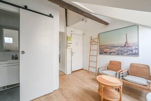 A compact living space with a kitchenette, a dining area with wicker furniture, and a cityscape painting on the wall.