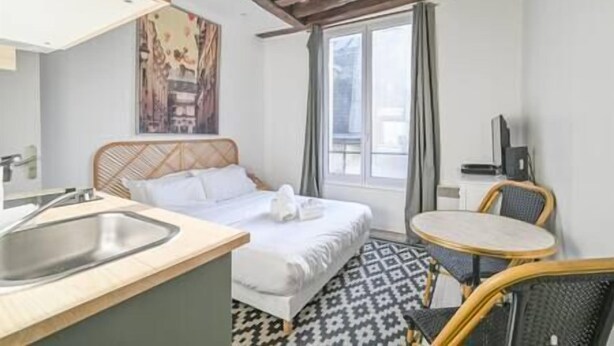 A hotel room with a bed, a small desk, a chair, a window, and a painting on the wall. - So cute typical french apartment (Paris)